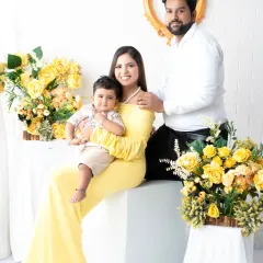 An elegant family portrait of three. The mother sits gracefully holding her son while the father stands beside them, creating a beautifully composed shot with a lovely yellow floral arrangement.
