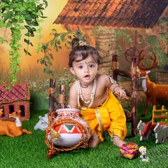 An expressive shot of a baby boy dressed as Krishna, surrounded by props like a pot of butter and toy deer. His curious look is what makes this themed photo so special.