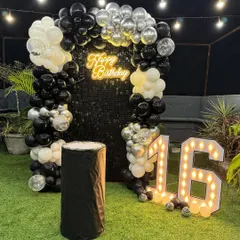 A sleek and modern 16th birthday setup on a terrace with an artificial grass floor. The black sequin shimmer wall is framed by a black, white, and silver balloon arch, complemented by large marquee numbers.