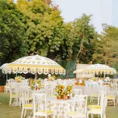 A classic daytime garden party setup. The use of traditional printed umbrellas, white furniture, and yellow floral centerpieces creates a cheerful and elegant atmosphere for a corporate brunch or luncheon.