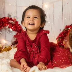 A huge, joyful smile lights up this Christmas-themed photoshoot. Dressed in a festive red dress, this little girl is surrounded by twinkling lights and holiday cheer.