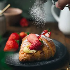 A moody, dramatic shot of a strawberry custard croissant being dusted with powdered sugar. The dark background makes the flaky pastry and bright red strawberries stand out beautifully.