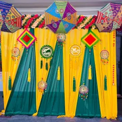 This is a classic fabric drape backdrop for a Haldi or Mehndi function. The combination of bright yellow and dark green curtains, accented with floral arrangements and traditional hangings, creates a vibrant and photogenic setting.