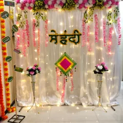 Another angle of the sober and sweet Mehndi setup. The combination of the bright lights behind the curtain and the colorful floral elements gives the space a warm and celebratory glow, perfect for a home function.