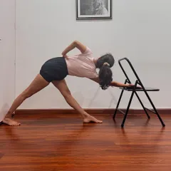 Parivrtta Trikonasana (Revolved Triangle Pose) is demonstrated here using a chair for support. The chair helps to maintain length in the spine and stability in the legs, allowing for a deeper and more refined twist.