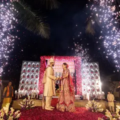 A moment of pure magic. The couple stands on a stage showered in flower petals, with fireworks lighting up the night sky behind them. This is the kind of grand, unforgettable varmala moment I love to create.