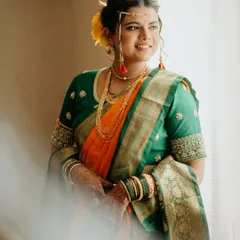 A candid shot capturing the bride's glow. Her hair is styled in a traditional bun adorned with a yellow flower, perfectly complementing her orange and green nauvari.