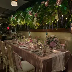 A full view of the enchanting birthday dinner setup for Anushka Sharma. A canopy of lush greenery and fairy lights hangs above the long table, which is adorned with elegant florals, candles, and sophisticated place settings.