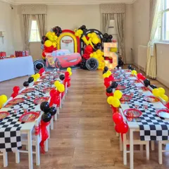 A complete room setup for a Disney Cars party. The long guest tables are decorated with checkered runners and themed tableware, all leading to the main backdrop with a giant Lightning McQueen.