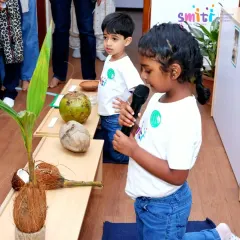 A young girl confidently speaks into a microphone, explaining the life cycle of a coconut to her family. Events like these build public speaking skills and self-assurance.