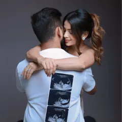 A playful and intimate announcement shot. The mom-to-be hugs her partner from behind, showcasing a strip of sonogram photos on his back, symbolizing their journey together.