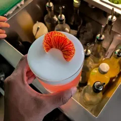 A perfectly fanned dehydrated orange slice sits atop a frothy sour. This type of intricate garnish requires skill and attention to detail, elevating the perceived value of the cocktail.