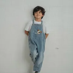 A simple and classic portrait of a young boy in denim overalls leaning against a white brick wall.