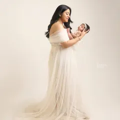 A full-length portrait of a mother in a flowing white gown, holding her newborn. It showcases the elegance and grace of new motherhood.