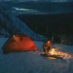 A camper starts a fire in the snow, the sparks flying up into the twilight. A glowing tent waits nearby. This image captures the essential warmth and light we create to thrive in the cold.