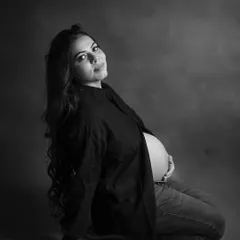 A relaxed and confident black and white portrait. The expecting mother is seated, wearing an open shirt over jeans, for a modern and stylish maternity look.