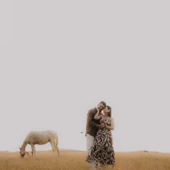 A wide, minimalist shot of a couple in a vast, golden field, with a horse grazing nearby. This image has a peaceful, storybook quality, emphasizing the couple's connection within a beautiful, expansive landscape.