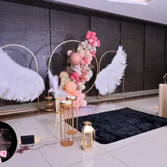 An elegant and serene baptism celebration. This design features large, feathered angel wings and a delicate balloon garland in shades of pink and peach, creating a beautiful photo backdrop. I use a mix of plinths and golden props to display the cake and other decor elements.