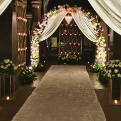 We designed this elegant entrance for a Roka ceremony. The walkway features a white carpet, a floral arch with pastel flowers and drapes, and candle-lit stands, creating a romantic and welcoming path.