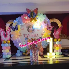 A complete stage setup for a butterfly-themed first birthday. It features a circular backdrop with the baby's name, a pastel balloon garland, large butterfly props, and an illuminated number '1'.