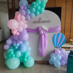 A modern and elegant naming ceremony decoration with a pastel balloon garland on a white circular backdrop. A small hot air balloon prop adds a whimsical touch.
