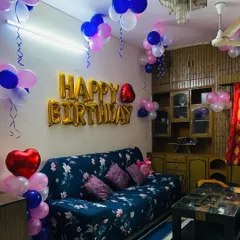 A cozy living room decorated for a birthday with pink, blue, and white balloons on the ceiling and a gold "Happy Birthday" foil banner.