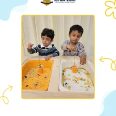 Two boys work side-by-side at the sensory table, using different tools to explore bins filled with rice and pasta. This parallel play is an important stage in social development.