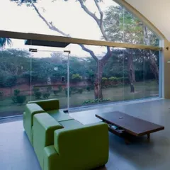 The interior of the repurposed Okhla shed feels open and connected to nature. The arched window frames a beautiful view of the trees outside, while the minimalist furniture and bright green sofa add a touch of modern comfort to the industrial space.