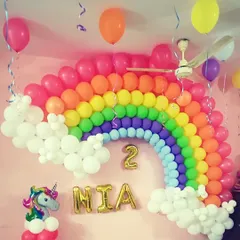 A beautiful rainbow balloon arch against a pink wall for a second birthday. This decoration includes balloon clouds, a unicorn balloon column, and the name "MIA" in gold foil letters below the rainbow.