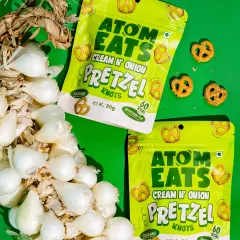 A vibrant shot of Atom Eats Cream 'n' Onion pretzels with a braid of fresh pearl onions against a bright green background. This composition creatively links the product to its core ingredient.