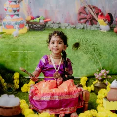 A smiling little Radha sits amidst a colorful Janmashtami setup. Her happy expression and the festive decorations make this a joyful and memorable shot.