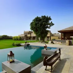 An infinity pool at a Chattarpur farmhouse designed to be a tranquil oasis. The clean lines of the Sadarhalli stone deck and the reflective water surface merge with the expansive lawn and distant trees, creating a serene landscape for relaxation and escape.