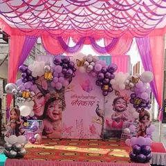 An elaborate stage setup for a traditional ceremony, likely an Annaprashan or baby welcome. The decoration includes a tent with pink and purple draping, a large backdrop with Krishna imagery, and extensive balloon work.