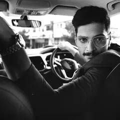 A candid, in-car portrait of actor Ali Fazal. This black and white shot creates a timeless, cinematic feel, capturing a moment of connection between the subject and the camera.