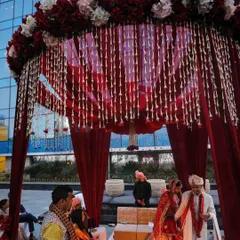 A classic round mandap design for a traditional Hindu wedding. The structure is draped in rich red fabric, with a canopy of red and white flowers and hanging tuberose strings.