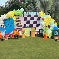 I created this action-packed car race theme for a 5th birthday party in a garden setting. The setup includes a checkered flag backdrop, custom racetrack cutouts, tire props, and a vibrant balloon arch in green, yellow, and blue.