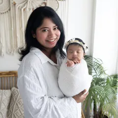 A new mother's joy is radiant in this photo. She is holding her sleeping, swaddled baby against the backdrop of my cozy, boho-inspired studio setup.