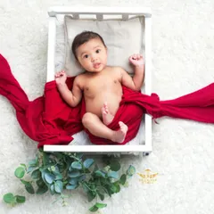 This setup features a miniature white bed prop, elegantly draped with a bold red fabric. It creates a striking visual contrast and a dramatic, fairytale-like portrait for this awake and curious baby.