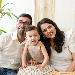 A classic family portrait with parents and their toddler smiling for the camera in a relaxed, indoor setting.