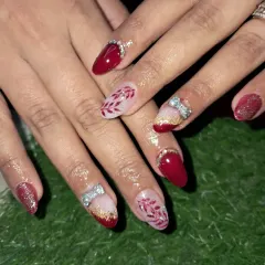 A close-up of a beautiful bridal nail set. We combined solid red nails, red glitter nails, and accent nails with delicate hand-painted leaves and 3D stone bows.