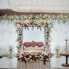 An elegant jhoola (swing) setup for a pre-wedding ceremony, framed by a beautiful arch of red and white flowers. The traditional brass lamp (samai) and golden pots add to the richness of the decor.