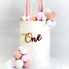 This 10-inch tall cake is a statement piece for a first birthday. The clean white design is accented with a boho rainbow and a cascade of pink and white edible spheres.