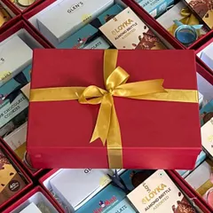 A vibrant red gift box, elegantly tied with a gold ribbon, stands out against a backdrop of dozens of identical hampers being prepared. This showcases our attention to detail on every single gift in a bulk order.