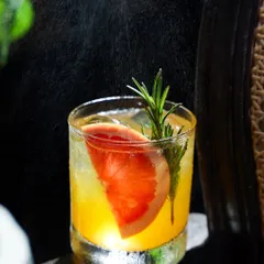 A close-up action shot of a grapefruit and rosemary cocktail being spritzed with zest. The backlighting catches the fine mist, adding a sensory dimension to the image that highlights the freshness of the ingredients.