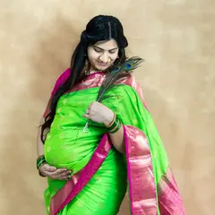 A creative studio portrait combining tradition with a touch of nature. The mom-to-be holds a peacock feather, which complements her green and pink saree perfectly.