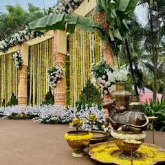 A beautiful setup for a traditional ceremony, featuring carved pillars, banana leaf decorations, and brass props like a Nandi statue and urlis filled with flowers.