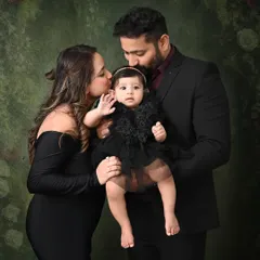 The elegance of family love. This portrait showcases a classic, timeless style with coordinated black outfits against a rich, dark green backdrop. The tender kisses for their one-year-old princess make the moment perfect.