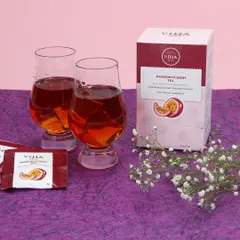 A lifestyle product photograph for Passion Fusion Tea. This setup includes the product box, individual tea sachets, and two glasses of the brewed beverage, arranged on a textured purple surface with delicate flowers.