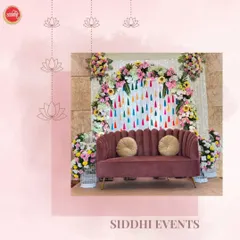 A chic and modern seating area for a celebration. A plush mauve sofa is set against a backdrop of colorful hanging tassels, framed by beautiful floral arrangements featuring sunflowers and roses, creating a perfect spot for photos.