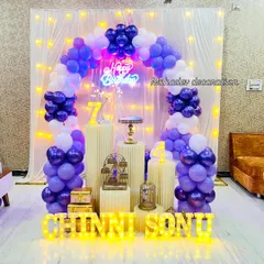 A vibrant birthday party setup for a 7th birthday, featuring a balloon arch in shades of purple and white. The backdrop is lit with fairy lights and includes a neon sign, multiple pedestals for the cake, and personalized marquee names.